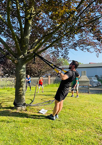 Mann in schwarzen Sportsachen zieht sich an TRX Baendern an einem Ast hoch